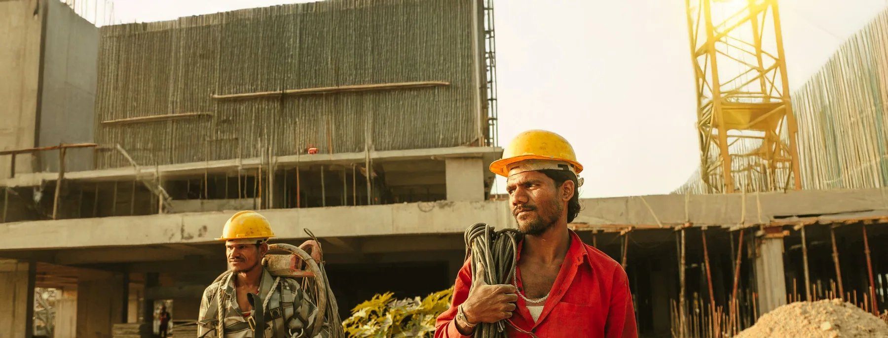 Two construction workers in hard hats carrying coils of cable across a building site